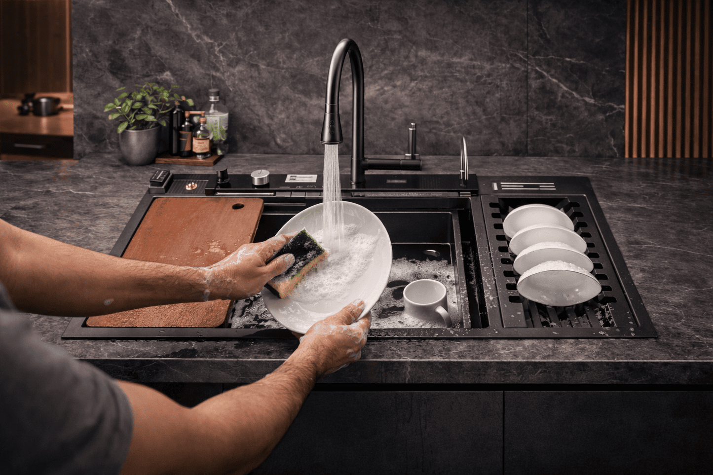 Person washing dishes in a modern kitchen sink with a dark countertop.